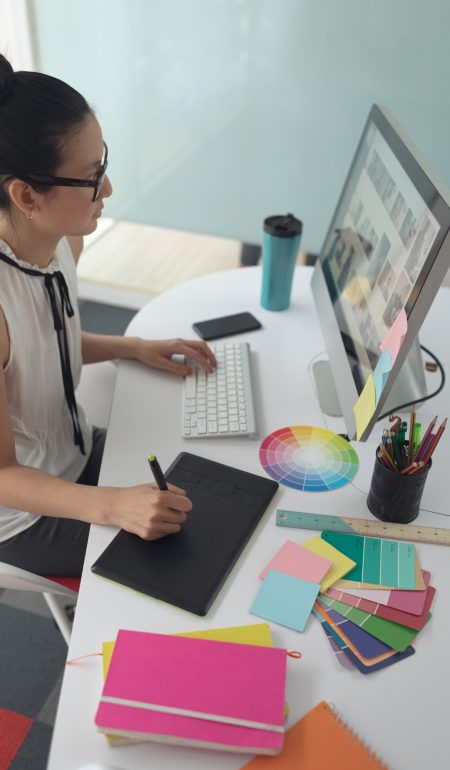 High angle view of Asian female graphic designer using graphic tablet at desk in a modern office